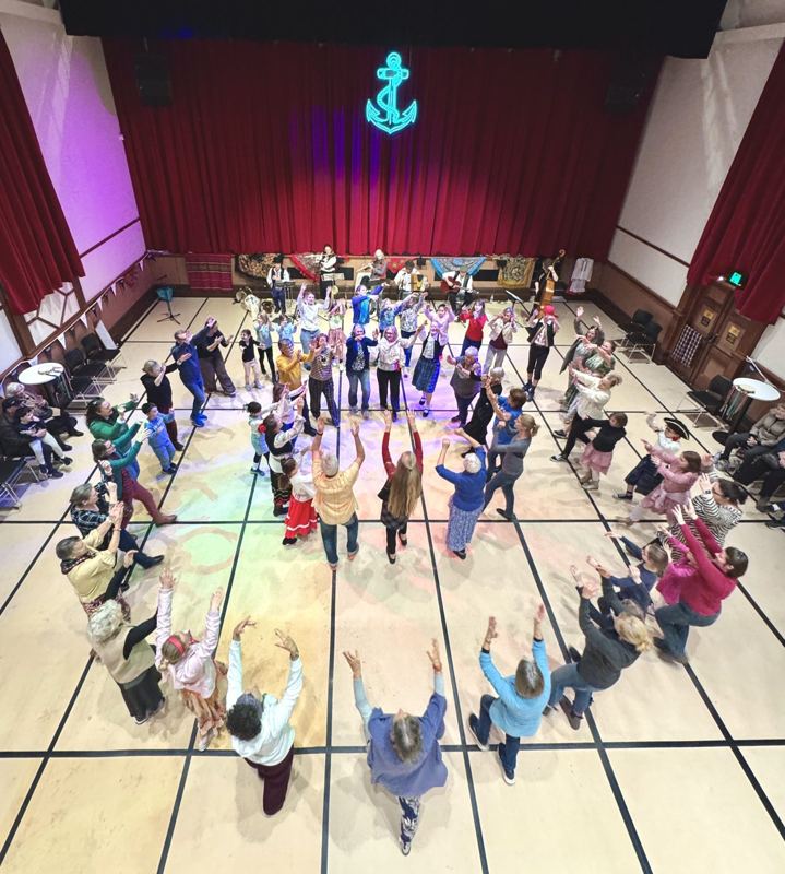 A packed hall of festival-goers dancing in a circle beneath a neon anchor